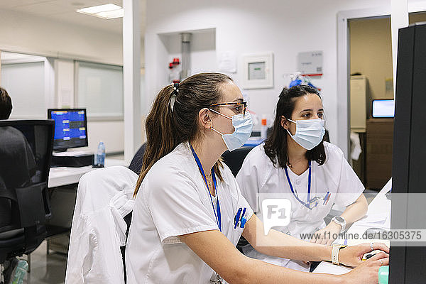 Female pharmacists wearing masks discussing over computer while sitting in hospital