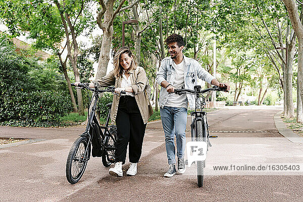 Lächelnder Freund und Freundin gehen mit Elektrofahrrädern auf der Straße