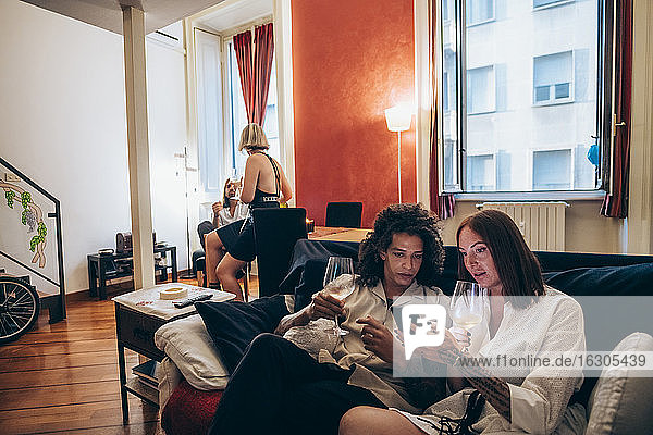 Frau zeigt einem männlichen Freund ihr Smartphone  während sie zu Hause auf dem Sofa Wein trinkt