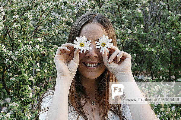 Smiling woman covering eyes with white flowers against plants in park during spring