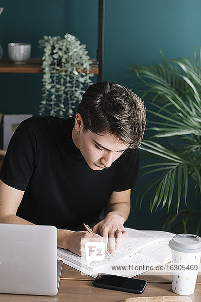 Junger männlicher Student  der in ein Buch schreibt  während er mit einem Laptop am Tisch sitzt und Hausaufgaben macht