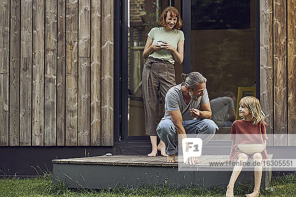Frau betrachtet Vater und Tochter  die vor einem kleinen Haus Kirschen essen