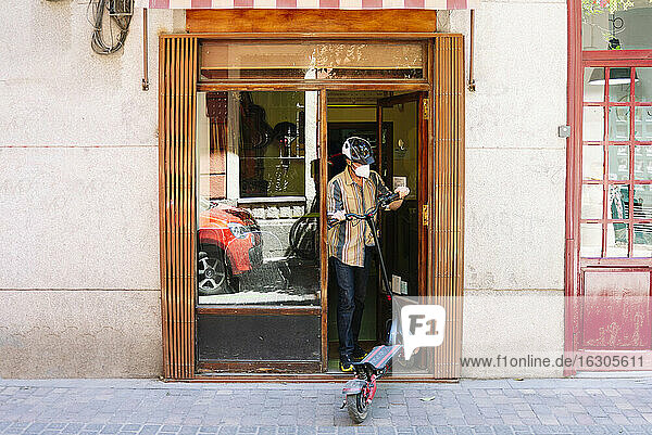 Älterer Mann mit Gesichtsmaske beim Entfernen eines Motorrollers auf der Straße beim Schließen der Werkstatt