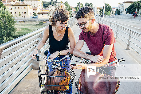 Mann zeigt seiner Freundin sein Smartphone  während er mit Fahrrädern auf einer Brücke steht