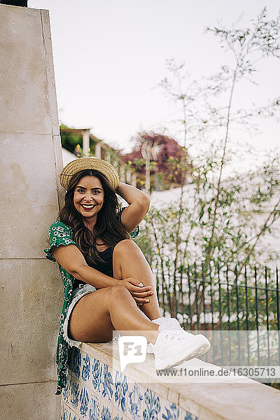 Lächelnde junge Frau mit Hut auf einer Stützmauer sitzend