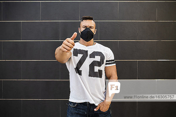 Macho-Mann mit Gesichtsmaske zeigt Daumen hoch  während er an der Wand steht