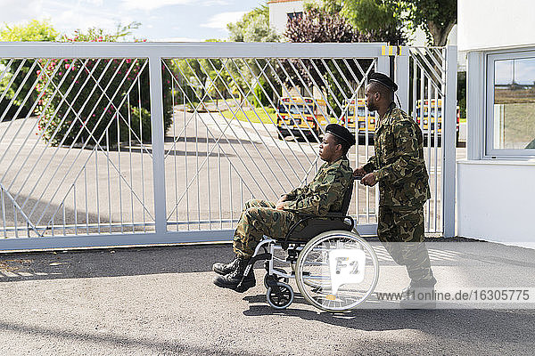 Army soldier pushing military soldier on wheelchair while standing against gate