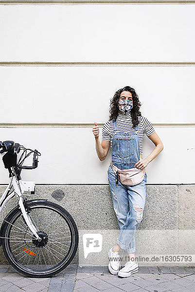 Mature woman wearing protective face mask showing thumbs up while standing by electric bicycle against wall