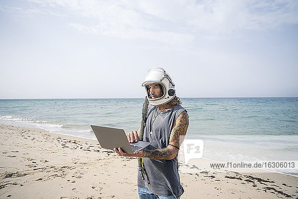 Mann mit Weltraumhelm  der einen Laptop benutzt  während er am Strand steht  Tarifa  Spanien