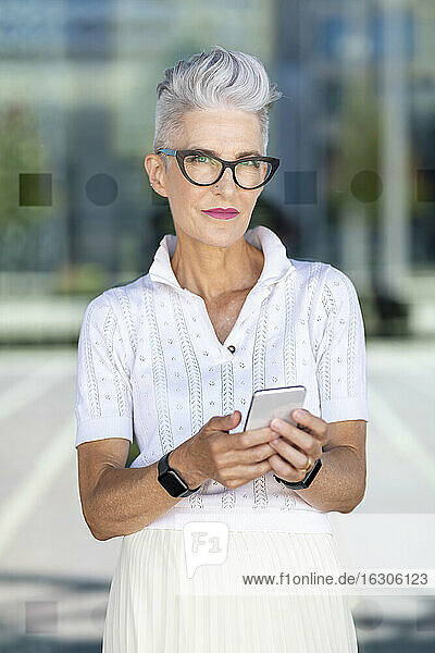 Lächelnde ältere Frau  die ein Smartphone benutzt  während sie vor einem Glasfenster steht