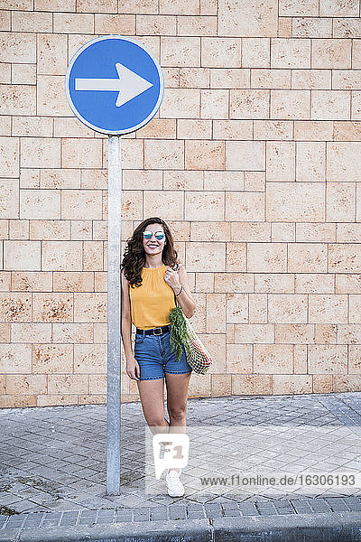 Lächelnde junge Frau  die sich auf ein Hinweisschild an einer Wand in der Stadt stützt