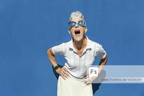 Frau mit offenem Mund und Hand auf der Hüfte steht gegen blaue Wand während sonnigen Tag