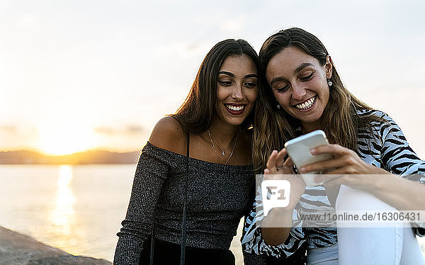 Junge Freunde telefonieren,  während sie bei Sonnenuntergang auf einer Stützmauer an der Promenade sitzen