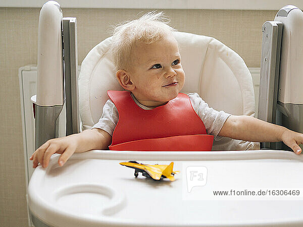 Lächelnder kleiner Junge sitzt mit einem Spielzeugflugzeug auf einem Hochstuhl und schaut in der Küche weg