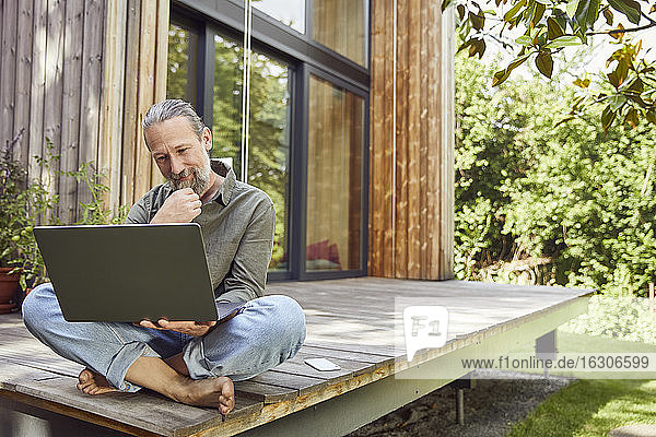 Bärtiger Mann benutzt Laptop  während er vor dem Haus sitzt