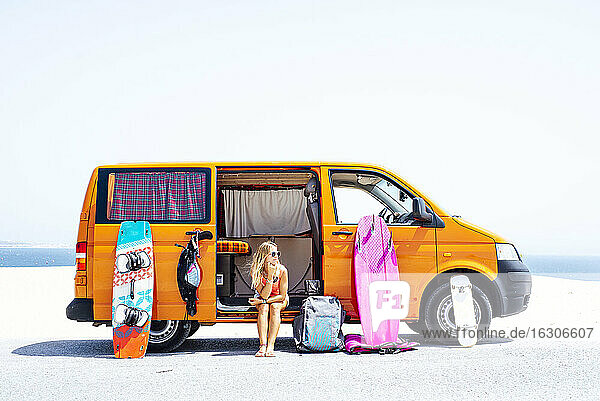 Frau sitzt mit der Hand auf dem Kinn gegen die Tür eines Lieferwagens am Strand an einem sonnigen Tag
