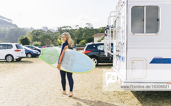 Frau  die an einem sonnigen Tag mit einem Surfbrett neben einem Wohnmobil spazieren geht