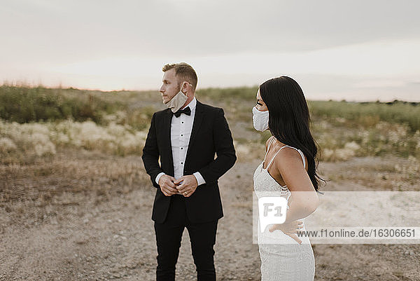 Braut mit Bräutigam  die einen Gesichtsschutz trägt  während sie im Feld während COVID-19 wegschaut