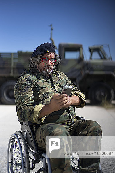 Armeesoldat  der im Rollstuhl sitzend an einem sonnigen Tag eine SMS auf seinem Smartphone schreibt