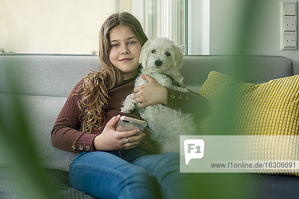 Lächelndes Mädchen  das ein Smartphone benutzt und einen Hund auf dem Sofa hält