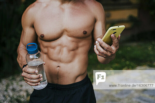 Nahaufnahme eines männlichen Sportlers ohne Hemd  der eine Wasserflasche hält und ein Smartphone im Garten benutzt