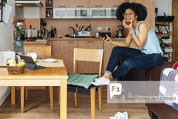 Female entrepreneur with curly hair using smart phone while eating bread at home
