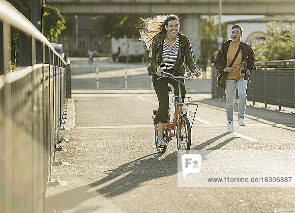 Boyfriend running behind happy girlfriend riding bicycle in city on sunny day