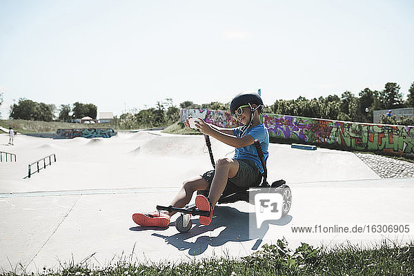 Boy taking selfie while sitting on hoverkart at skateboard park against sky during sunny day
