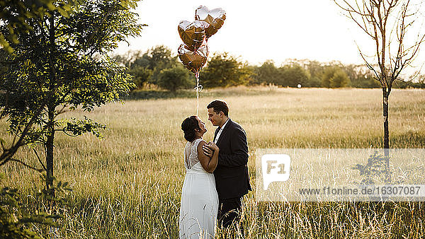 Brautpaar hält Heliumballons in Herzform  während sie in einem Feld bei Sonnenuntergang stehen