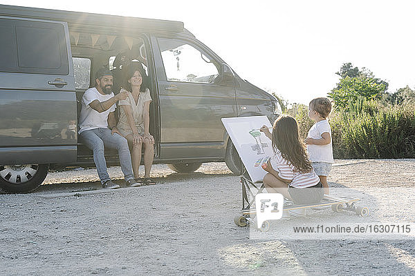 Glückliche Eltern sitzen im Van  während die Tochter bei Sonnenuntergang von ihrer Schwester gemalt wird