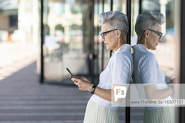 Reflektierendes Bild einer älteren Frau  die sich an ein Glasfenster lehnt  während sie ein Smartphone in der Stadt benutzt