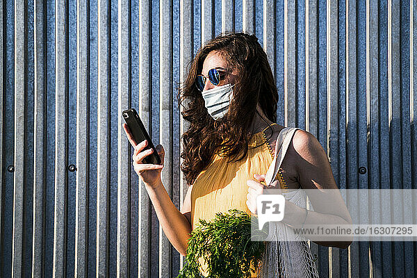 Junge Frau mit Gesichtsmaske  die mit Handy und wiederverwendbarer Netztasche an einer Metallwand steht