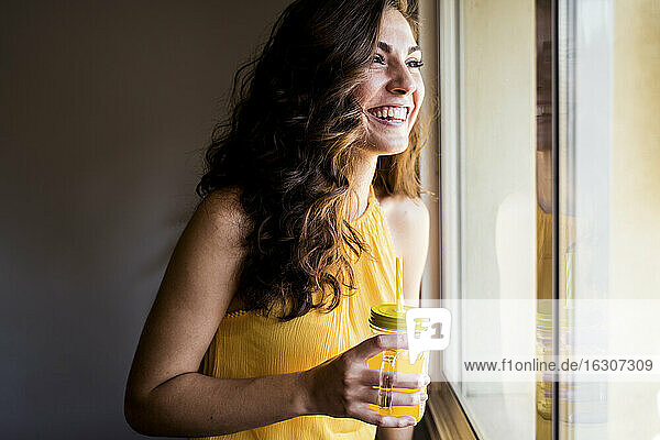 Glückliche Frau  die ein Einmachglas mit Saft in der Hand hält  während sie durch das Fenster zu Hause wegschaut