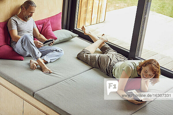 Mann benutzt digitales Tablet  während Frau auf dem Bett zu Hause ein Buch liest