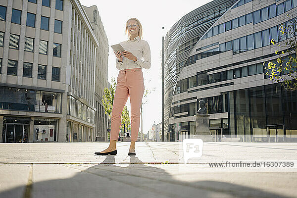 Smiling female professional holding digital tablet while standing on footpath in city during sunny day