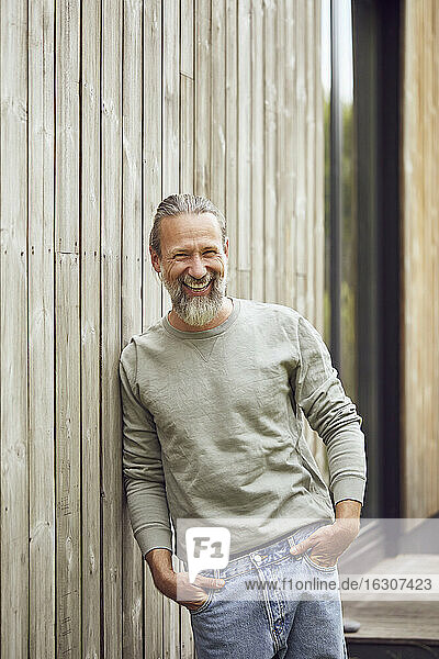 Happy bearded mature man with hands in pockets standing by wall at yard