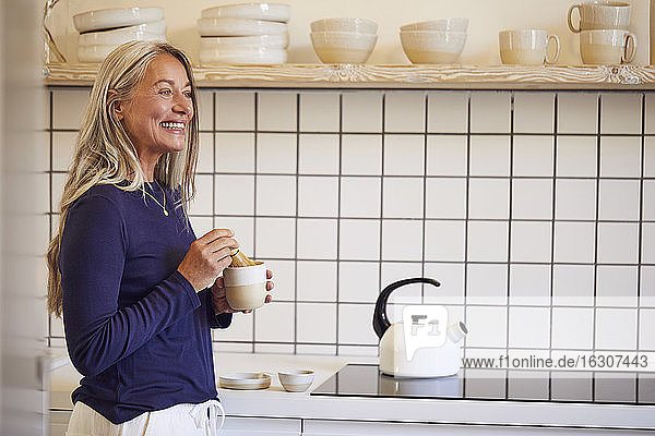 Lächelnde Frau trinkt Tee  während sie in der Küche steht
