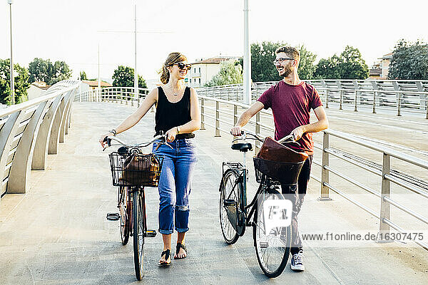 Glückliches Paar  das sich unterhält  während es mit Fahrrädern auf einer Brücke gegen den klaren Himmel läuft