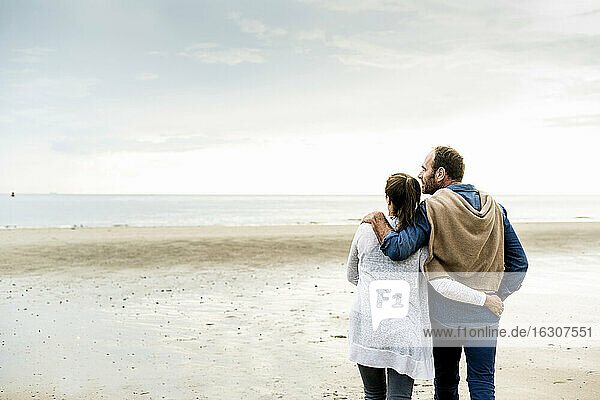 Couple with arms around looking at sea against cloudy sky during weekend