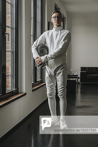 Young fencer with foil and mask standing by windows in loft at home