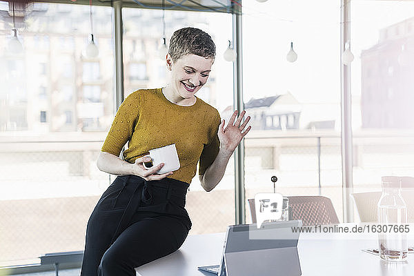 Lächelnde Geschäftsfrau sitzt auf einem Schreibtisch im Büro und führt eine Videokonferenz durch