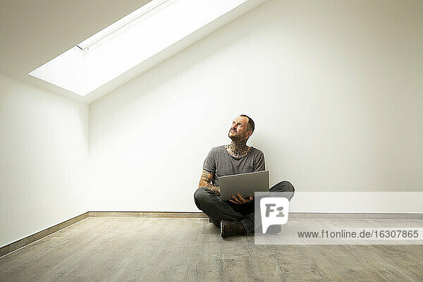 Mature man with laptop looking through window while sitting against white wall in attic of new house