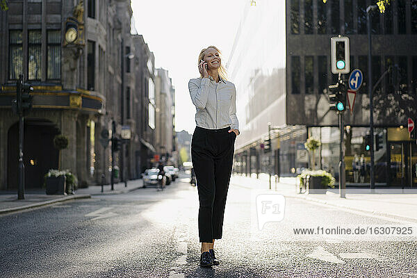Smiling businesswoman talking through mobile phone while looking away and walking on street in city