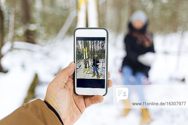 Man photographing girlfriend by skis through smart phone in forest during winter