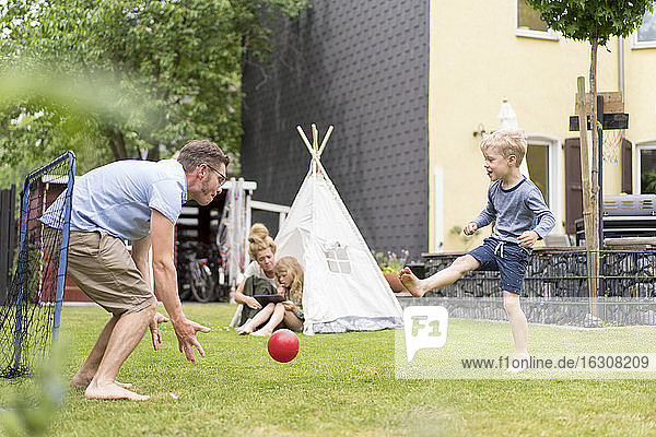 Mann und Sohn spielen Fußball  während die Frau mit ihrer Tochter im Zelt im Hinterhof sitzt