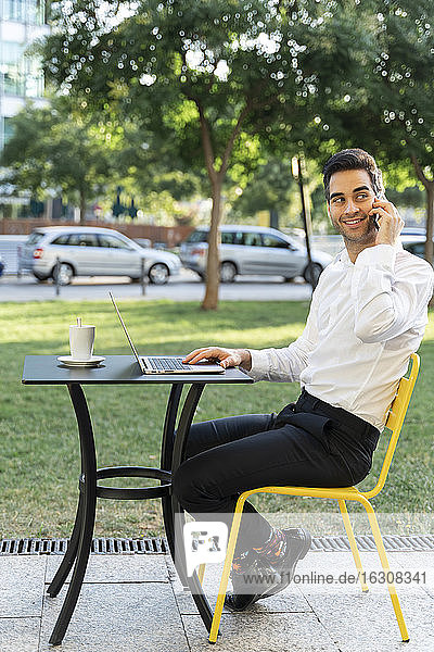 Gutaussehender Geschäftsmann mit Laptop auf dem Tisch  der in einem Café im Freien über sein Handy spricht