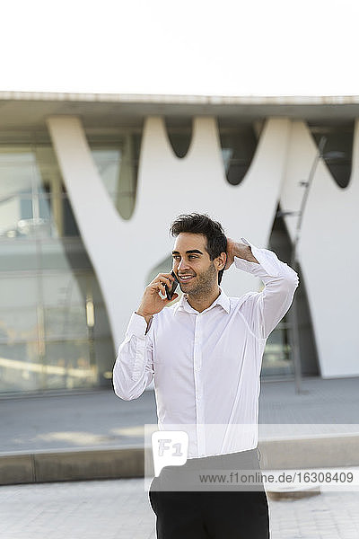 Gutaussehender Geschäftsmann mit Hand im Haar  der über ein Smartphone spricht  während er vor einem Gebäude steht