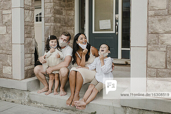 Lächelnde Familie mit Kindern  die einen Gesichtsschutz tragen  während sie auf den Stufen vor dem Haus sitzen