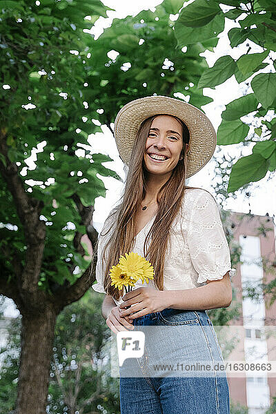 Lächelnde junge Frau mit Hut und gelben Blumen in der Hand im Park stehend