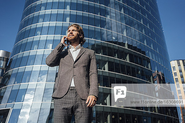Geschäftsmann schaut weg  während er mit dem Handy vor einem modernen Bürogebäude an einem sonnigen Tag spricht
